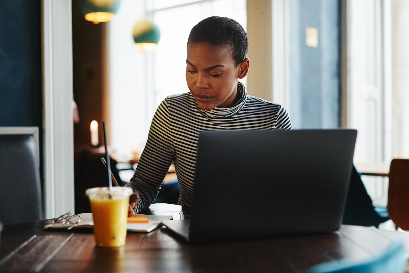 Young African American female entrepreneur sitting in a cafe while writing down notes and working on a laptop