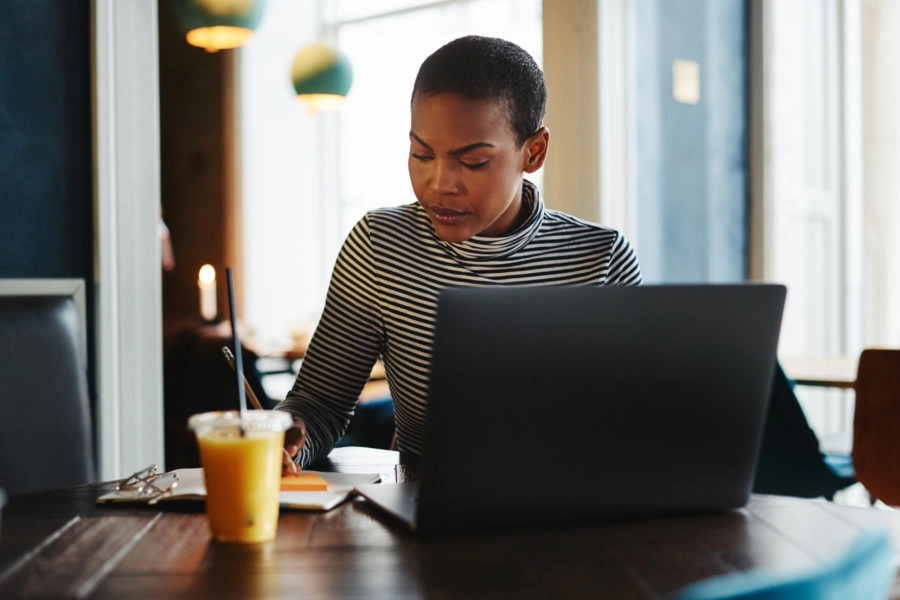 Young African American female entrepreneur sitting in a cafe while writing down notes and working on a laptop