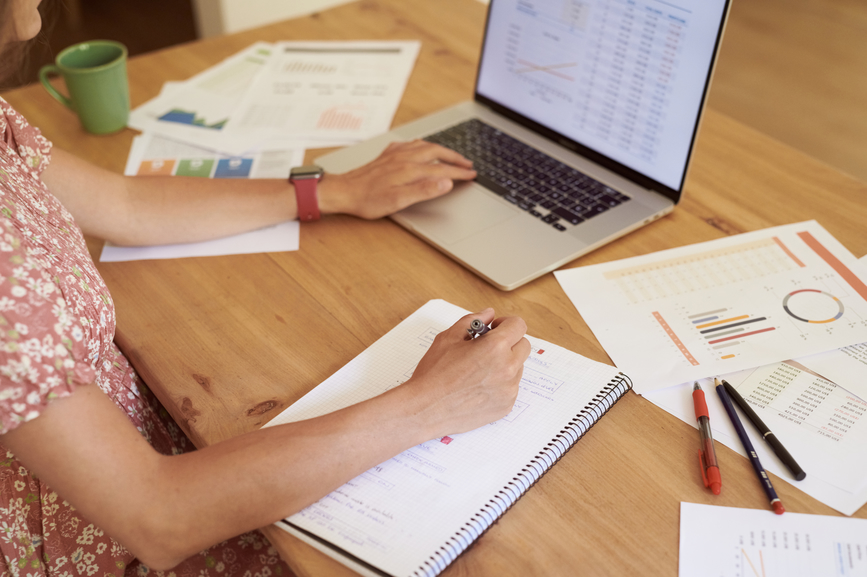 Freelancer making notes and working on laptop at wooden desk with scattered documents and stationery.