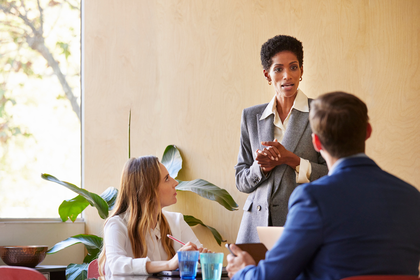 African American woman in gray suit clasping hands and speaking to attentive managers during business meeting in light office in daytime.